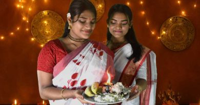Two young Bengali girls dressed in traditional sarees hold a decorated thali with a lit diya, flowers, and fruits during Diwali celebrations. The background features golden plates, string lights, and a small deity statue, creating a festive and spiritual ambiance.